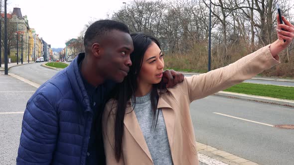 A Black Man and a Young Asian Woman Take a Selfie with a Smartphone in a Street in an Urban Area