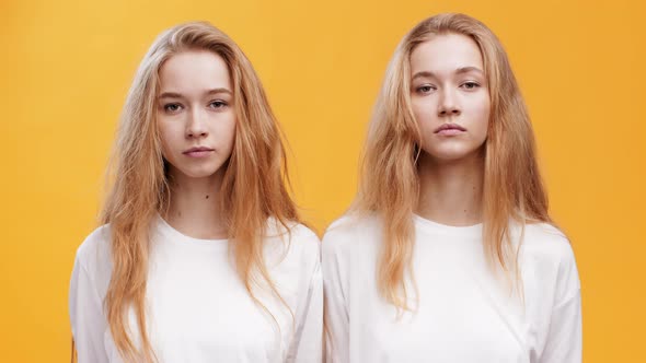 Portrait of Two Serious Twin Sisters Looking at Camera Posing Over Orange Background