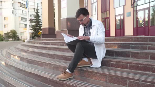 A Medical Student Sits on the Stairs Outside the University and Studies the Medical Report Preparing