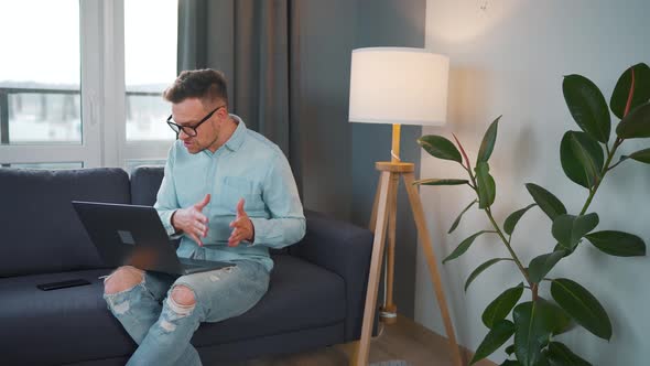 Casual Dressed Man with Glasses Sitting on the Couch and Using Laptop to Make a Video Call From Home