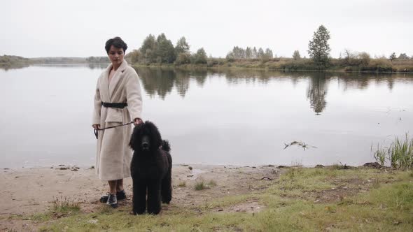 An Elegant Young Brunette in a White Coat Stands By the River and Holds a Large Black Poodle on a