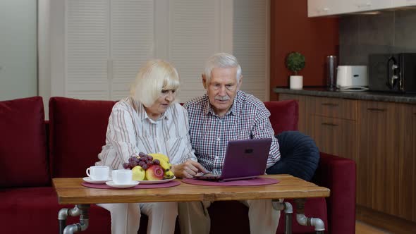 Senior Pensioner Couple with Digital Laptop Pc Computer at Home. Resting on Sofa in Cozy Living Room