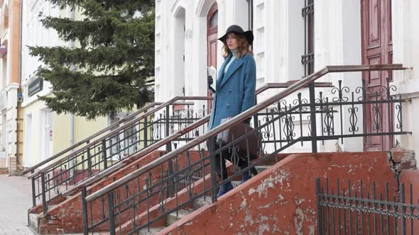 Young Redhaired Woman in a Blue Coat and Black Hat with a Suitcase on the Background of the Old City