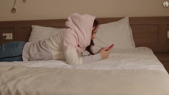 Young Woman in Hood Lying on Bed with Mobile Phone