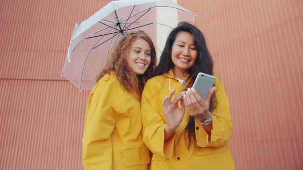 Cheerful Young Women in Raincoats Using Smartphone Outdoors Under Umbrella