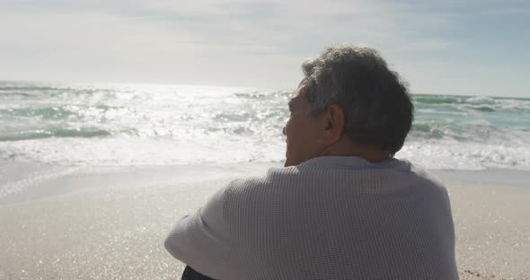 Back view of hispanic senior man sitting on beach at sunset