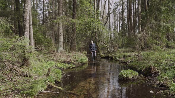 Young Man in Rubber Boots Walks Along the Bed of a Clear Stream in the Forest