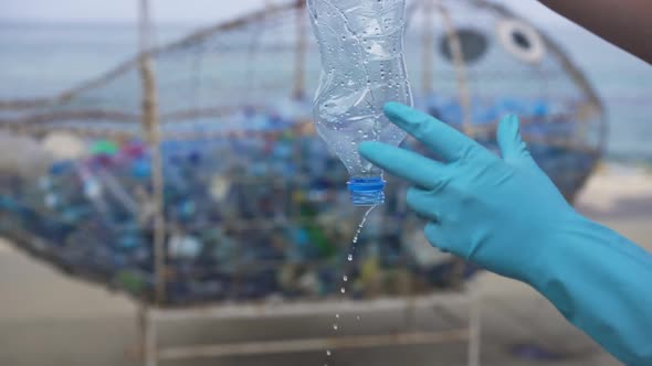 Female Caucasian Hands in Gloves Crumpling Empty Plastic Bottle with Trash Collected at Background