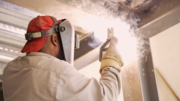 Man in red cap, welding helmet, safety gloves welding metal structure together