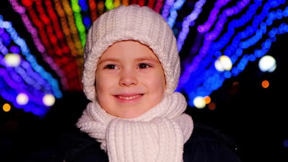 A Handsome Child in a White Hat Smiles Standing Under Christmas Garlands in the Evening