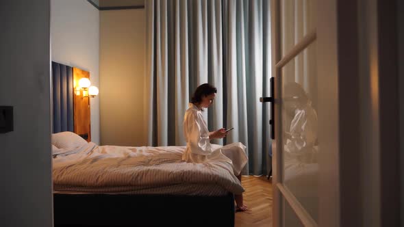 Cheerful Woman in Pajamas Typing on the Phone on the Bed in the Bedroom at Home