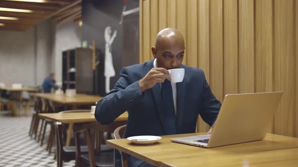 African Male Freelancer Using Laptop While Working Remotely and Drinking Coffee in Cafe