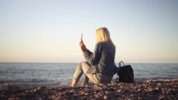 a woman sits on the seashore and writes a sea sunset on her phone.