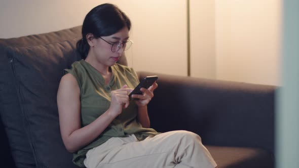Asian woman using smartphone checking social media while sitting on a sofa in the living room.