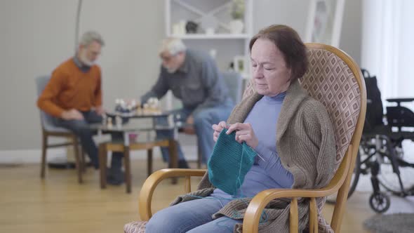 Old Caucasian Woman Knitting in Armchair As Blurred Men Playing Chess at the Background. Elderly