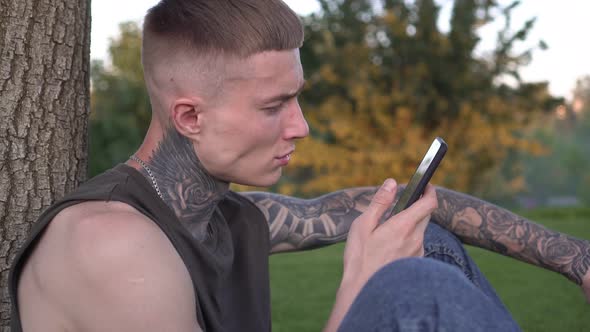 Young Modern Man with Tattoos Uses Smartphone Outdoors in the Park