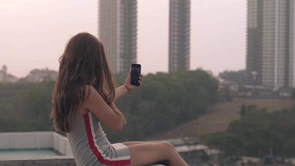 Brunette Sits on Roof and Takes Selfie with Black Smartphone