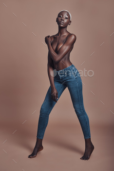 g African woman looking at camera and covering breast with hand while standing against brown background