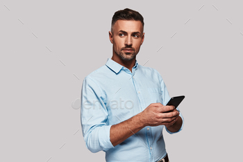  smart casual wear using his smart phone and smiling while standing against grey background