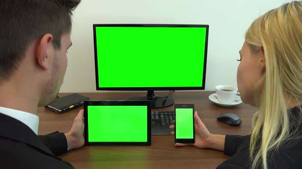 Two office workers sit at a desk and look at a computer, a tablet and a smartphone with green screen