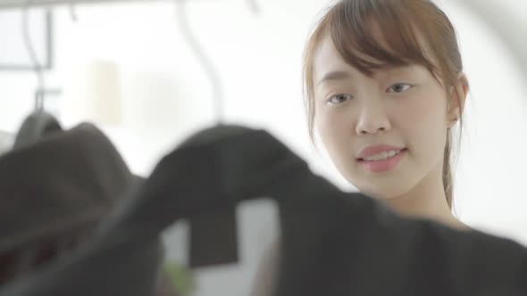 Beautiful young asian woman choosing clothes from rack with hangers at room.