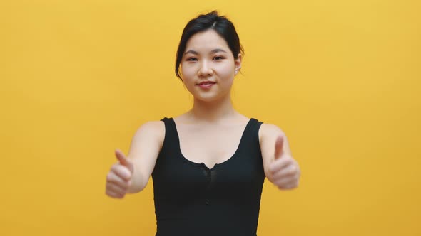 Beautiful Young Asian Woman Gesturing with Hands, Thumbs Up Isolated on Yellow Background