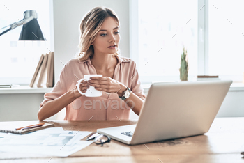 sual wear holding a cup of coffee and looking at laptop while sitting in modern office