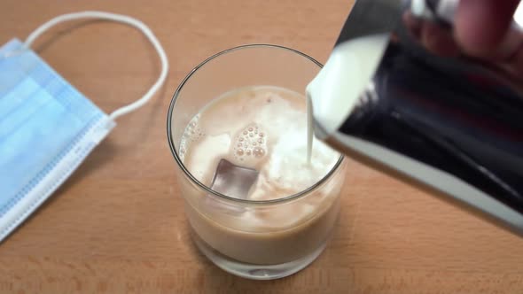 Milk is pouring from a metal jug into a glass with cold tea and ice cubes on a wooden cafe table