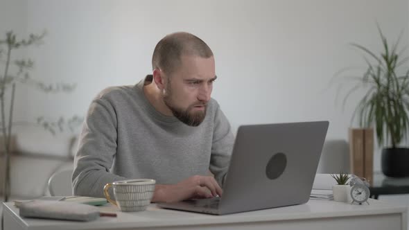 Angry man with beard in a gray jacket working and reading bad news on a laptop at home