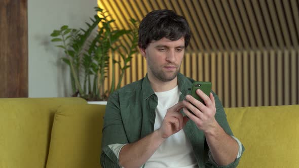 Smiling Young Man Holding Mobile Phone While Sitting on Yellow Sofa at Home and Celebrating