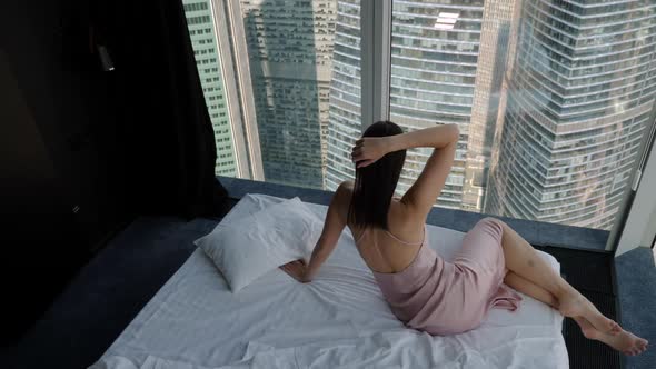 Woman in a Nightgown Lies on a Bed Near a Window in a Corner in a Skyscraper