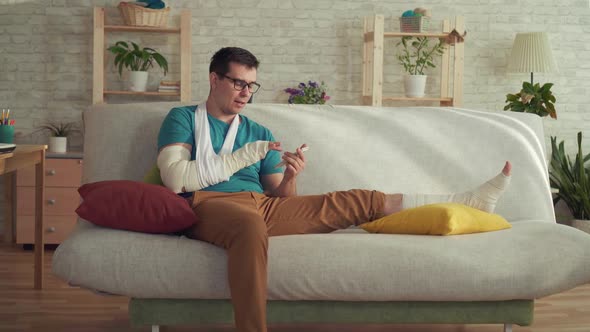 Young Man with a Broken Arm and Leg Sitting on a Sofa and Using a Smartphone