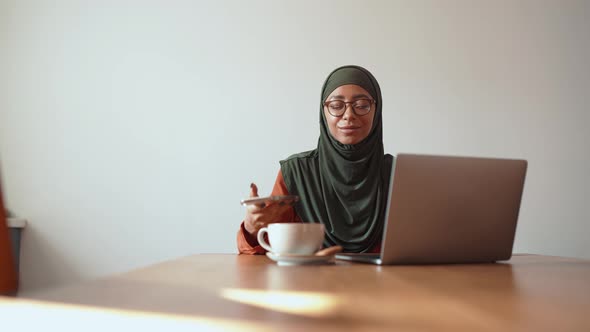 Positive Muslim woman wearing eyeglasses working on laptop and looking at phone
