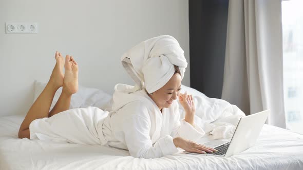 Cheerful Asian Woman Lying in Bed After a Shower in a Bathrobe Using a Laptop Communicates Via Video