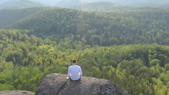 Drone Footage of a Freelancer Working on a Laptop in the Open Air Sitting on the Edge of a Rock