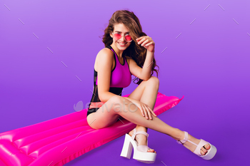 asses on purple background. She wears colorful swimsuit sitting on pink air mattress.