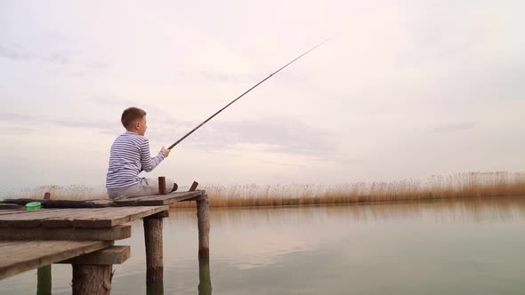 A Boy Sits on a Wooden Bridge and Catches a Fish on Fishing Rod