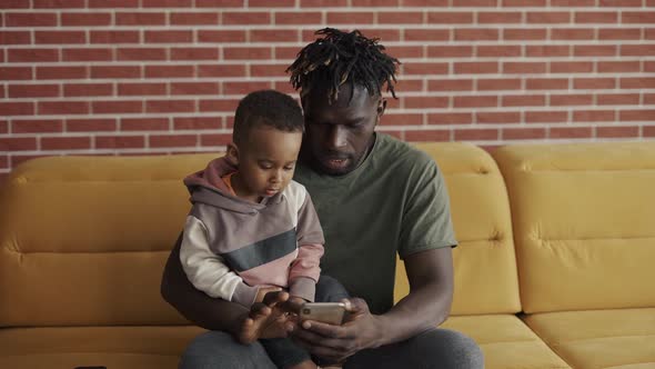 African American Father Sitting with Son on Sofa Using Smartphone