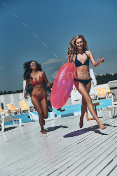 iful young women in bikini running outdoors together while one of them carrying inflatable ring