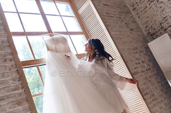 hrobe holding her wedding dress and smiling while standing near the window