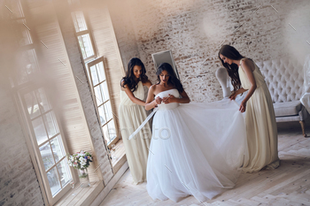 tractive young women adjusting a wedding dress on a bride while standing near the window