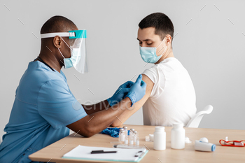 f population against coronavirus. Adult african american doctor in protective mask giving injection to patient in clinic, with equipment on table
