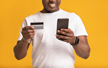 on. Cheerful millennial african american man in white t-shirt uses smartphone and credit card, isolated on orange background, studio shot, cropped