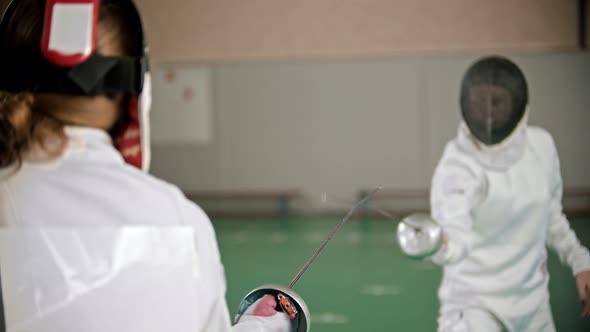 Two Young Women in Protective Costumes at Fencing Training in the School Gym - One Woman Attacks and