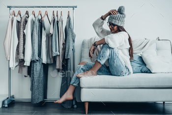 in casual wear adjusting her hat and looking away while sitting on the couch at home near her clothes hanging on the racks