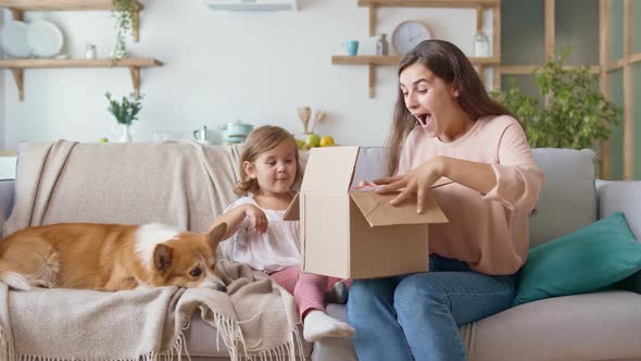 Happy Mother and Daughter Unpacking the Order. A Young Girl Takes From the Carton Box a Red Present.