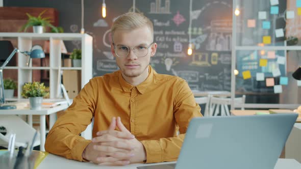 Portrait of Blond Young Man Looking at Camera Sitting at Desk with Laptop