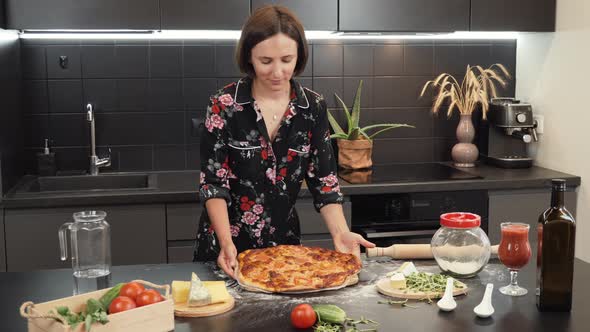 Young cheerful woman puts tasty baked pizza on wooden table. Process of making pizza