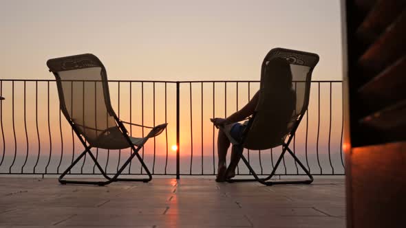 Young Woman Sits in a Sun Lounger on the Balcony with a Beautiful View of the Sea and Mountains