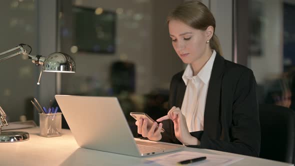 Young Businesswoman Scrolling on Smartphone in Office at Night